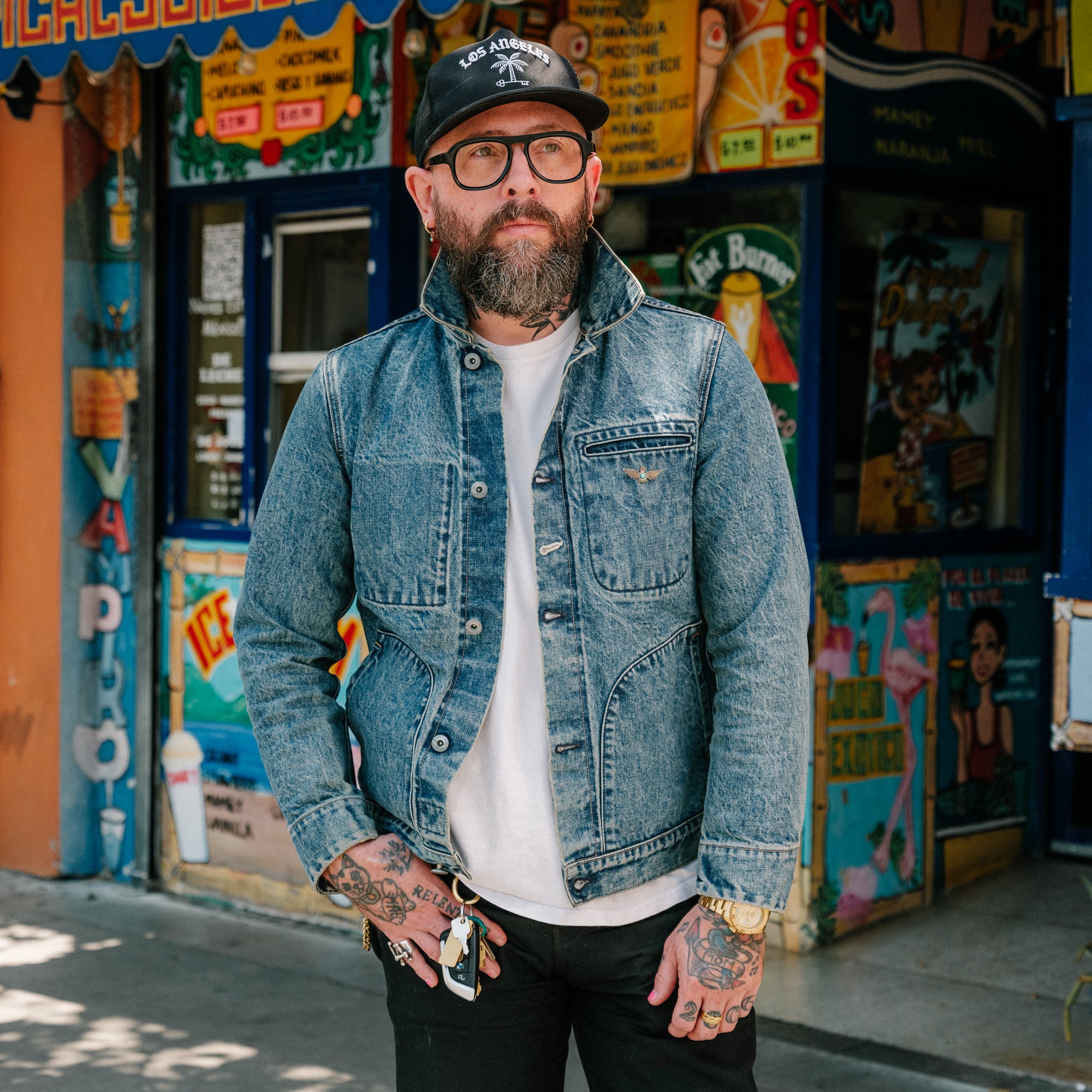 Man with tattoos wearing a denim jacket and cap in front of a colorful store.