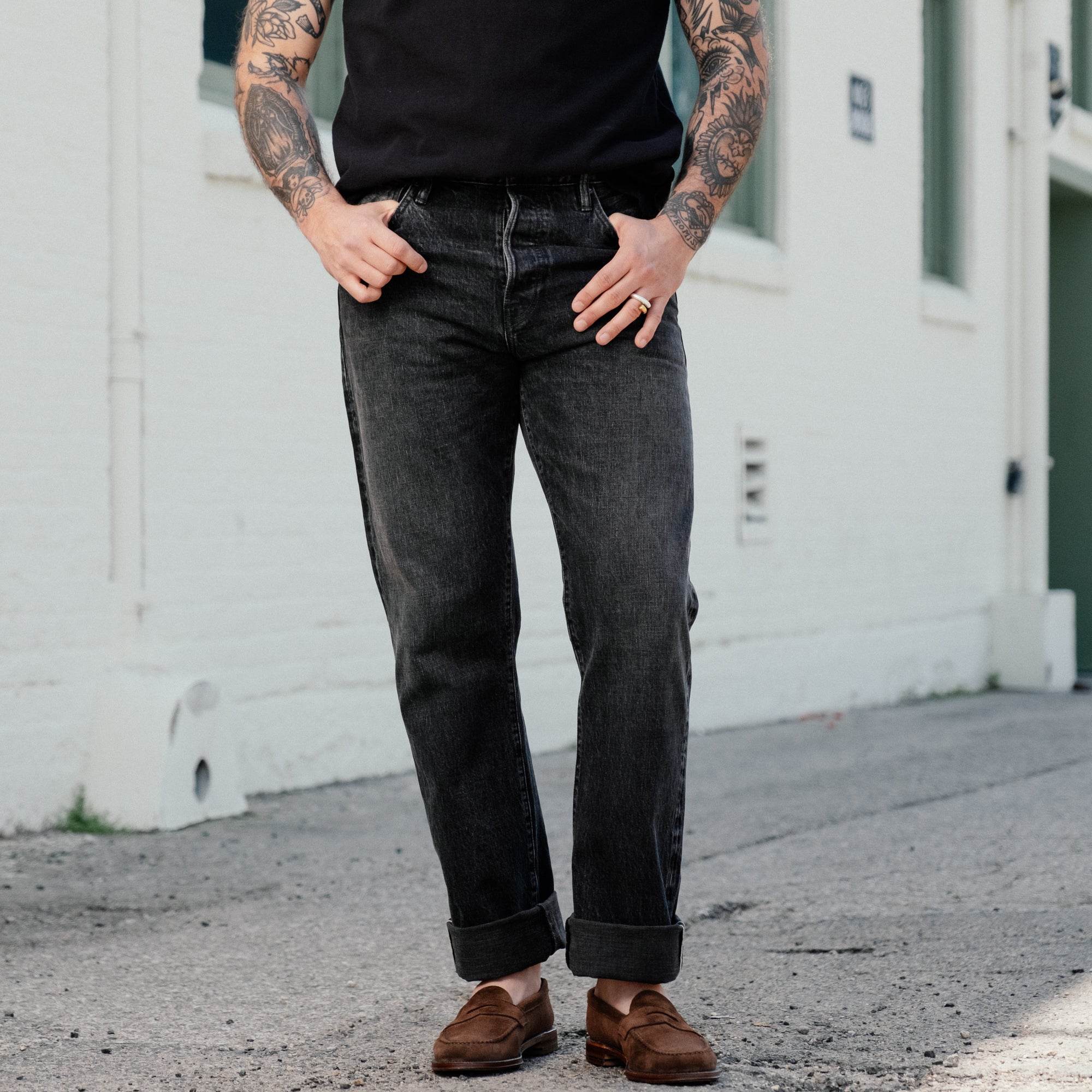 A person wearing dark charcoal gray straight-leg jeans with rolled cuffs, paired with a black t-shirt and brown suede loafers, standing outdoors against a white brick wall.