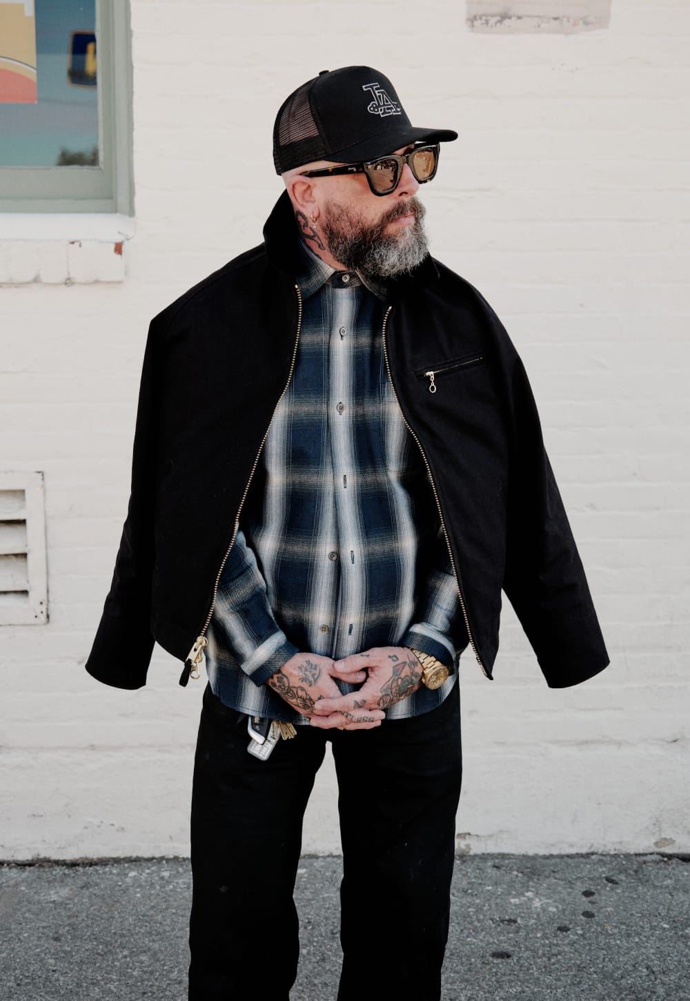 A man wearing a black zip-up jacket over a blue and white plaid flannel shirt, black pants, and a black trucker hat, standing against a white brick wall.
