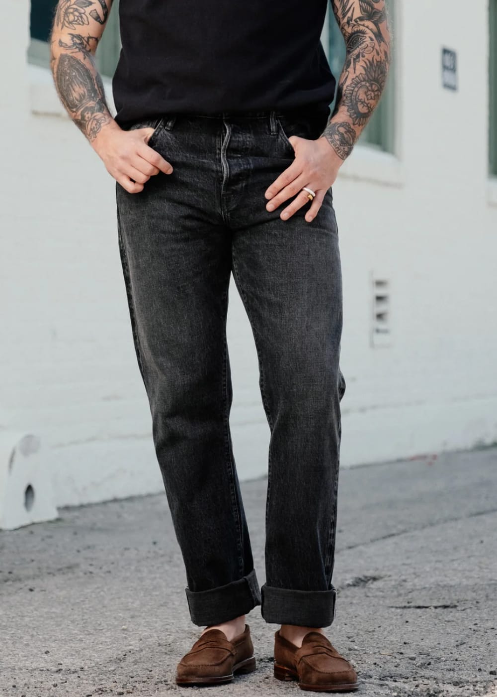 A person wearing dark charcoal gray straight-leg jeans with rolled cuffs, paired with a black t-shirt and brown suede loafers, standing outdoors against a white brick wall.