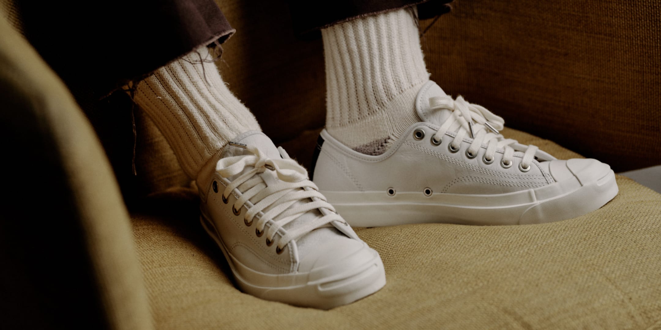 A pair of low-top white canvas sneakers with white laces and white ribbed socks, shown on a person sitting on a textured beige couch.