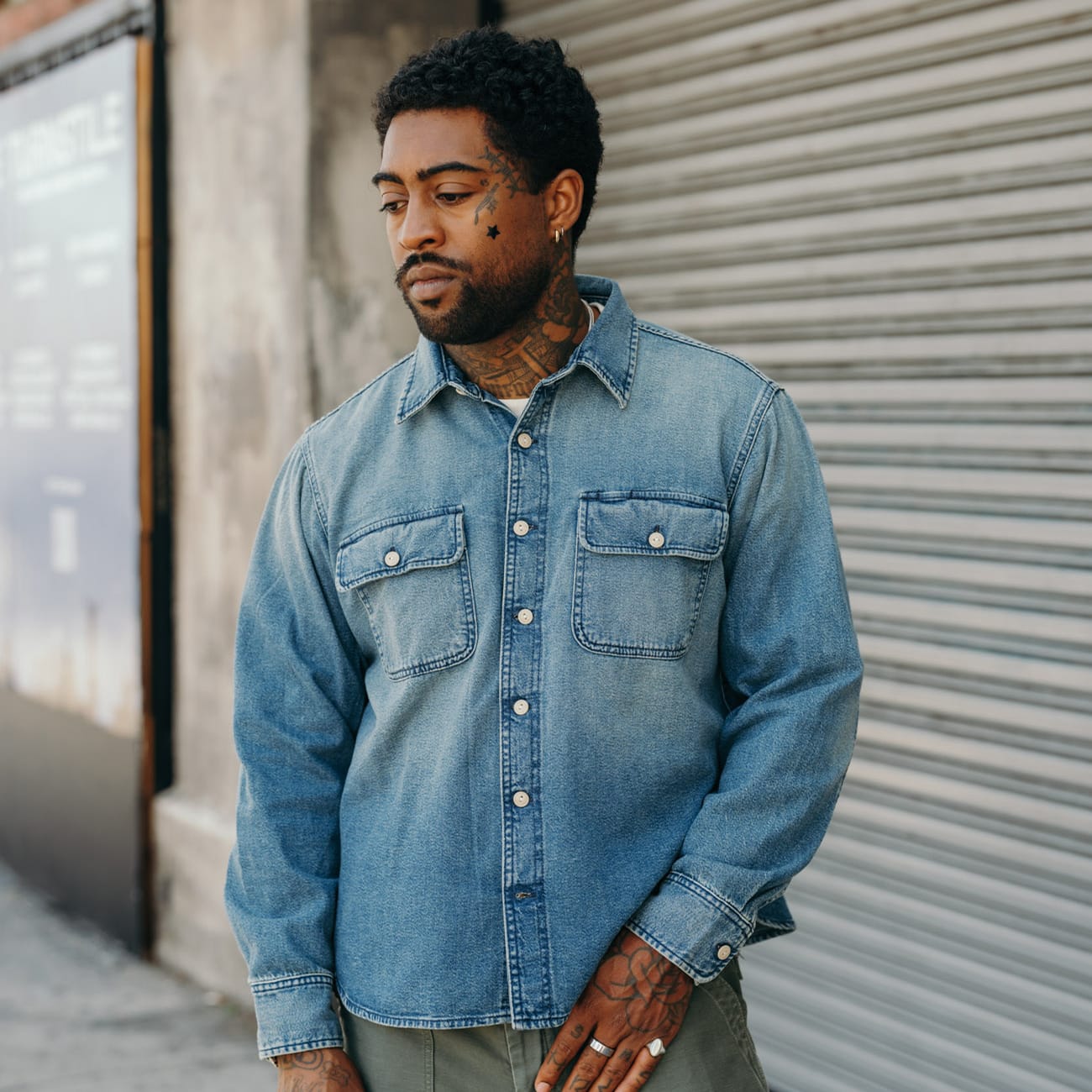 A man wearing a light blue denim button-up shirt with two chest pockets, standing in front of a closed metal garage door.